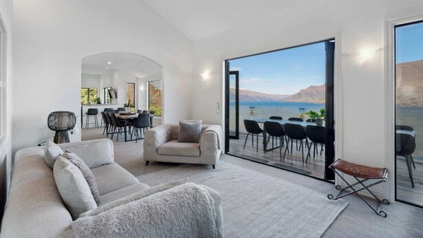A modern living room with a sofa, armchair, and a bench, a dining area with chairs and a table, and a large sliding glass door leading to a balcony with a view of mountains and a body of water. - Million Dollar Views | Central Luxury | Private Hot Tub (Queenstown)