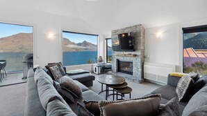 A modern living room with a stone fireplace, large windows, and a view of a lake and mountains. - Million Dollar Views | Central Luxury | Private Hot Tub (Queenstown)