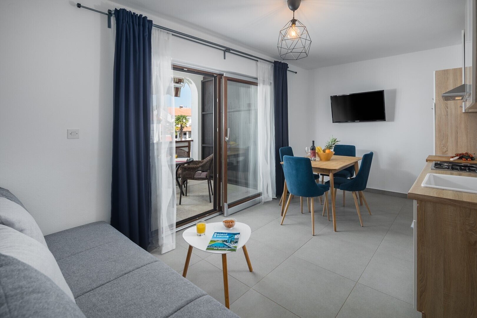 A modern living room with a sofa, a dining table with chairs, a kitchen area with a TV, and a sliding glass door leading to an outdoor area.