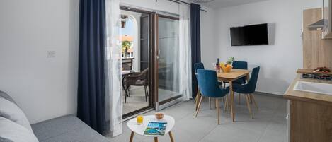 A modern living room with a sofa, a dining table with chairs, a kitchen area with a TV, and a sliding glass door leading to an outdoor area.