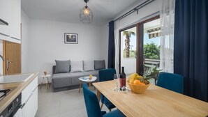 Eine moderne Küche mit Holzarbeitsplatte, ein Essbereich mit Holztisch und blauen Stühlen, ein graues Sofa mit Kissen und ein großes Fenster mit Vorhängen.