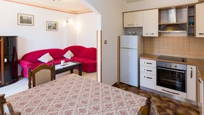 Eine kleine Küche mit Kühlschrank, Backofen und Schränken. Ein Essbereich mit Tisch und Stühlen ist zu sehen, und im Hintergrund steht ein rotes Sofa.