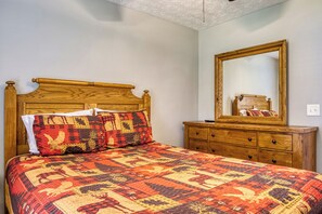 Una cama con cabecera de madera, colcha estampada y dos almohadas. Se observa un aparador de madera con cajones y un espejo encima.