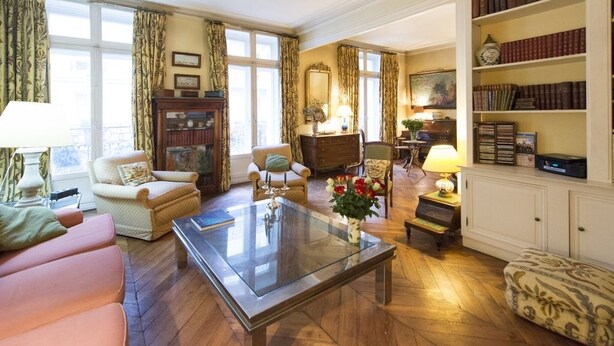 A spacious living room with a glass coffee table, comfortable seating, a bookshelf, and large windows with curtains.