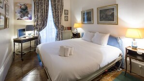A hotel room with a large bed, a desk with a computer, a chair, and a lamp. There are two paintings on the wall and a window with curtains.