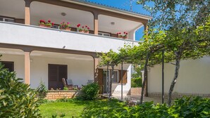 Un edificio de dos plantas con un balcón en el piso superior, una puerta en la planta baja y un jardín con plantas y un árbol.