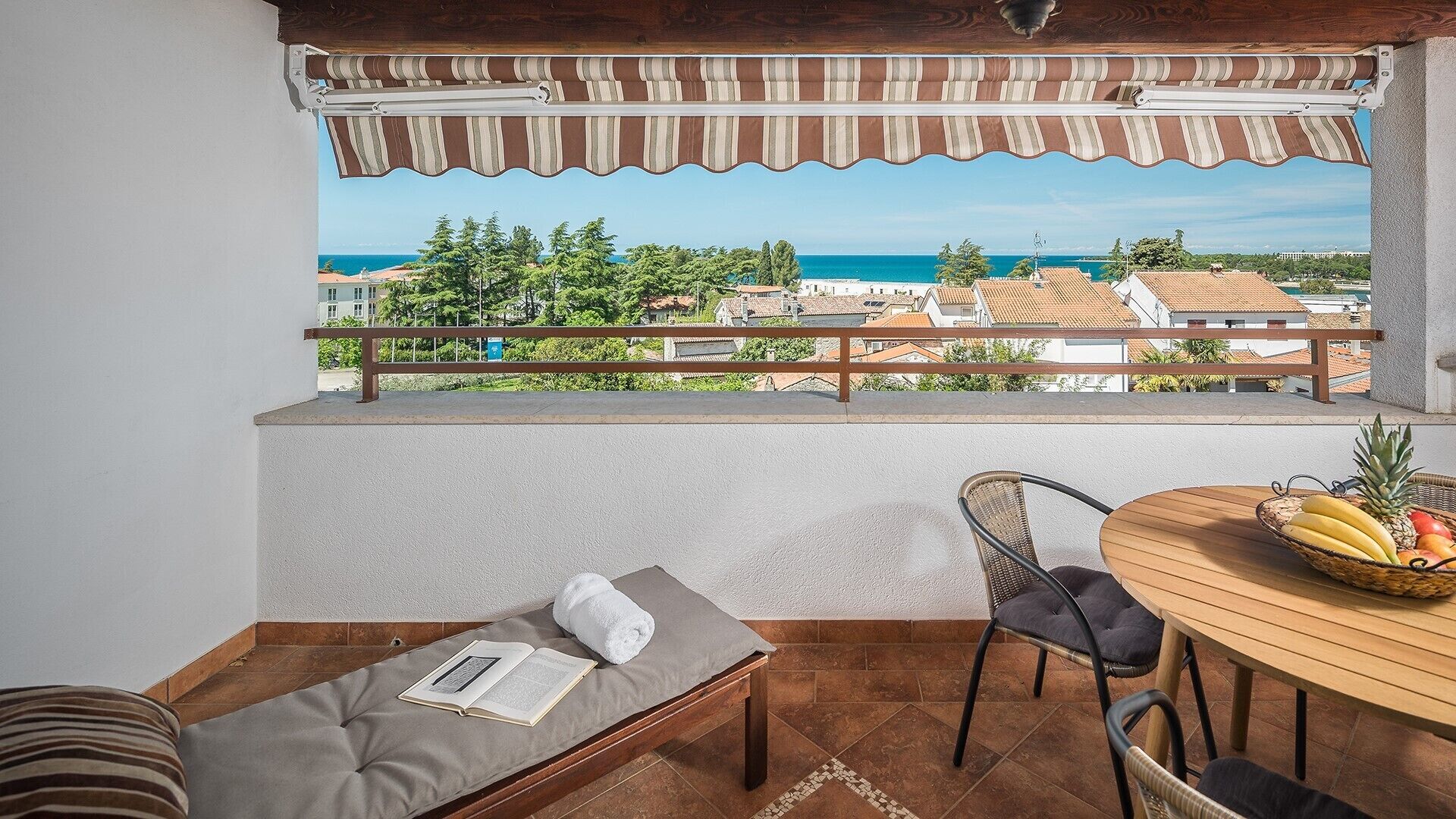 A balcony with a view of the sea, a wooden table with fruit, a cushioned bench, and a wicker chair.