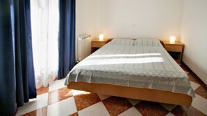 A bedroom with a bed, two bedside tables with lamps, a radiator, and a window with curtains.