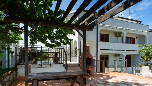 An outdoor patio with a wooden table and benches, a brick oven, and a pergola with climbing plants.