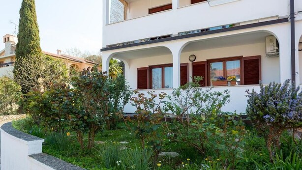 Un bâtiment à deux étages doté d’un balcon, entouré d’un jardin où poussent différentes plantes et fleurs.
