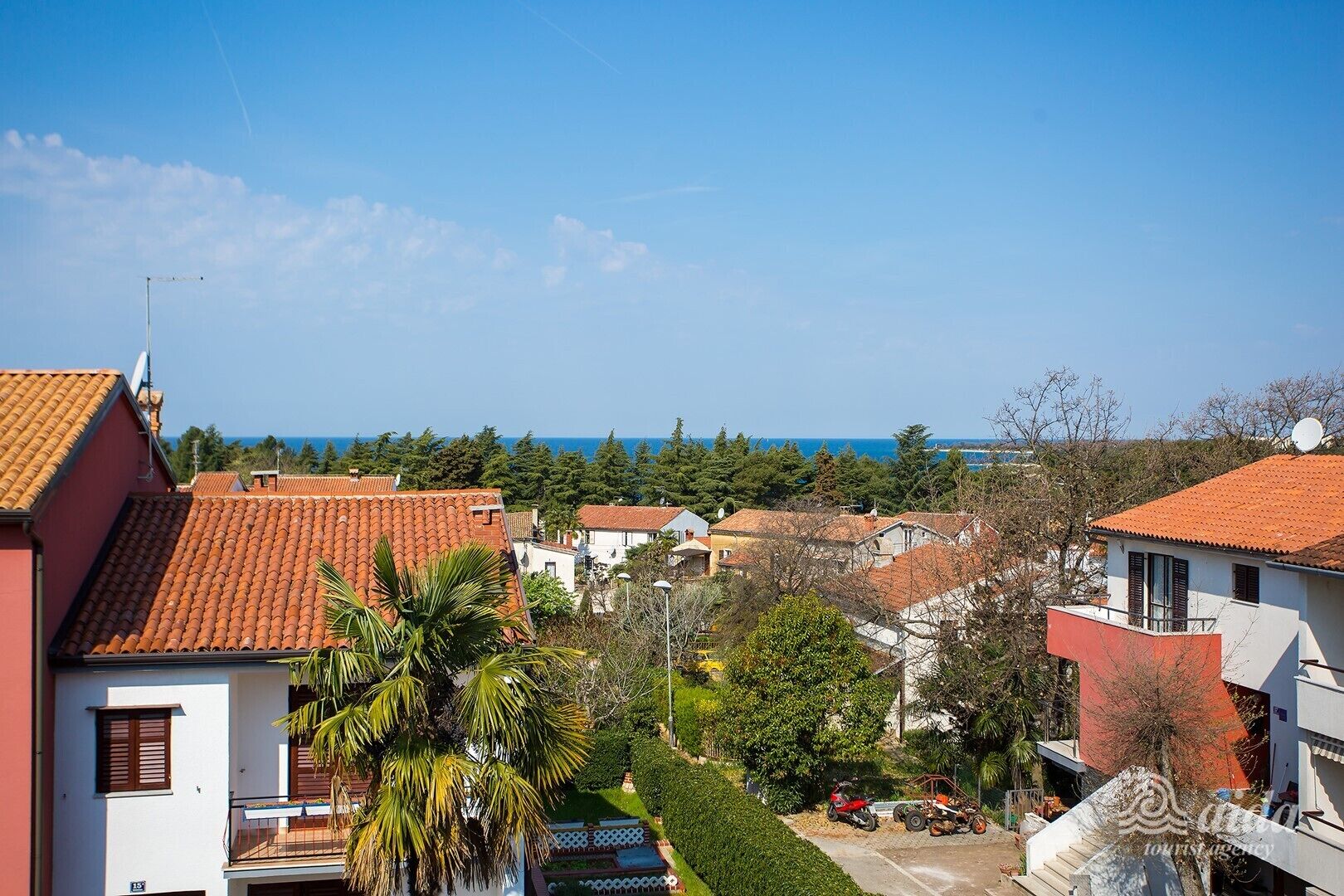 A coastal residential area with terracotta-tiled roofs, a clear blue sky, and a view of the sea.