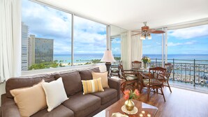 Un salon moderne doté d’une grande fenêtre donnant sur une plage et une marina, et aménagé avec un canapé en cuir marron, une table à manger avec des chaises et un ventilateur de plafond.