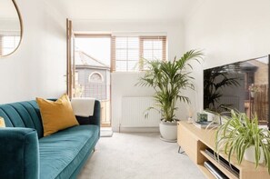 Una sala de estar con un sofá color turquesa, una planta grande en maceta, un televisor sobre un mueble de madera y una ventana con persianas.