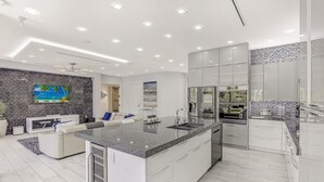 Una cocina moderna con una isla central, electrodomésticos integrados y una zona de estar con sofá blanco y televisor de pantalla plana.