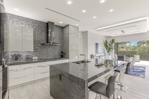 A modern kitchen with a granite countertop, stainless steel appliances, and a large island with bar seating. - Modern Home | Outdoor Kitchen & Steps to Beach (Marco Island)