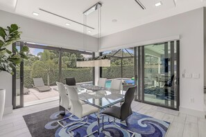 A modern dining area with a glass table and chairs, large windows, and a view of the outdoors. - Modern Home | Outdoor Kitchen & Steps to Beach (Marco Island)