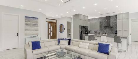 A modern living room with a sectional sofa, a glass coffee table, and a kitchen area with a built-in stove and cabinets.