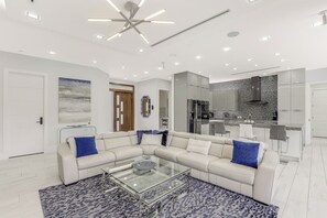 A modern living room with a sectional sofa, a glass coffee table, and a kitchen area with a built-in stove and cabinets.