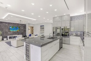 A modern kitchen with a central island, built-in appliances, and a seating area with a white sofa and a flat-screen TV. - Modern Home | Outdoor Kitchen & Steps to Beach (Marco Island)