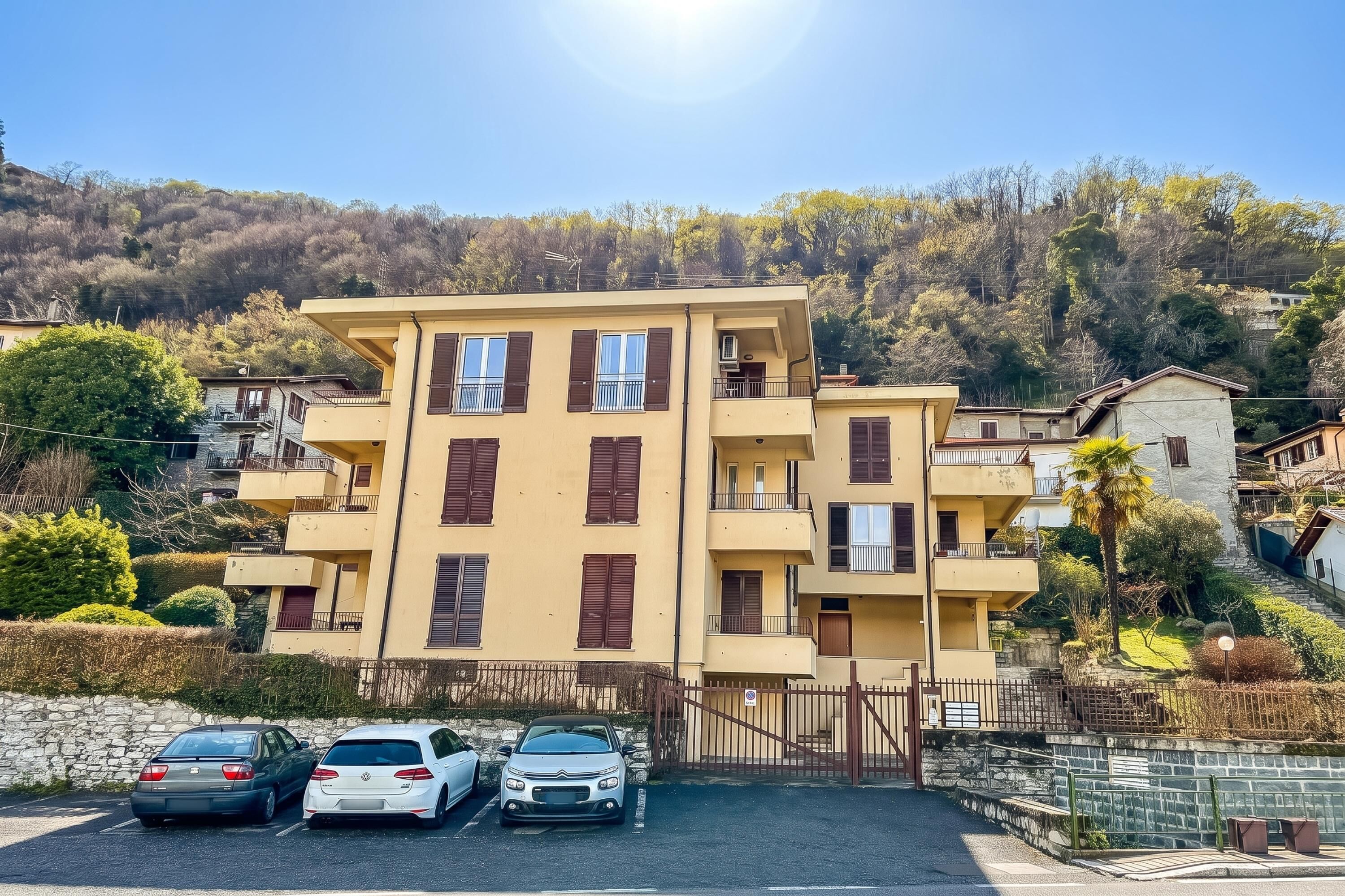 A multi-story residential building with balconies, a parking area with three cars, and a hillside with trees in the background.