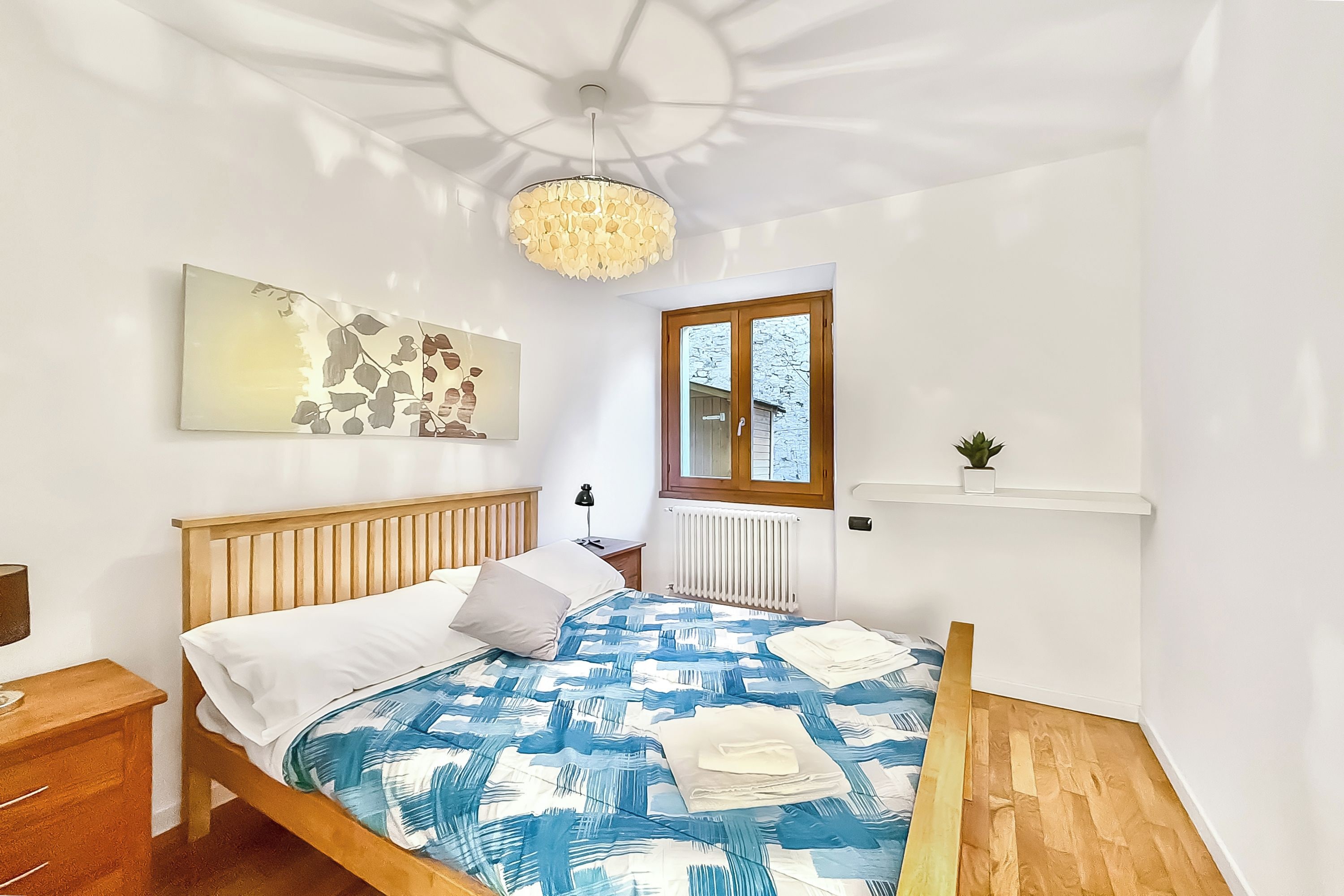 Une chambre avec un lit en bois, une couvre-lit à motifs bleus et blancs, un mur blanc orné d’un tableau, une table de chevet en bois, une lampe sur pied, une fenêtre donnant sur l’extérieur et un luminaire au plafond.