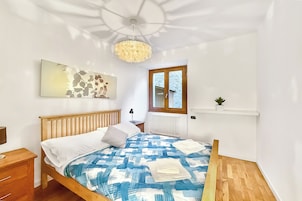 Un dormitorio con una cama de madera, una colcha azul y blanca estampada, una pared blanca con un cuadro, una mesita de noche de madera, una lámpara de pie, una ventana con vistas al exterior y un accesorio de iluminación en el techo.