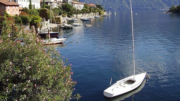 A calm lake with a sailboat docked near the shore, surrounded by buildings and mountains in the background. - Regina Grande (Laglio)