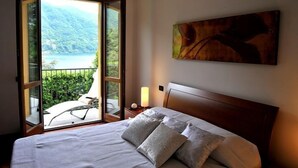A bedroom with a bed, a nightstand, a painting, and a view of a balcony with a mountain and lake. - Regina Grande (Laglio)