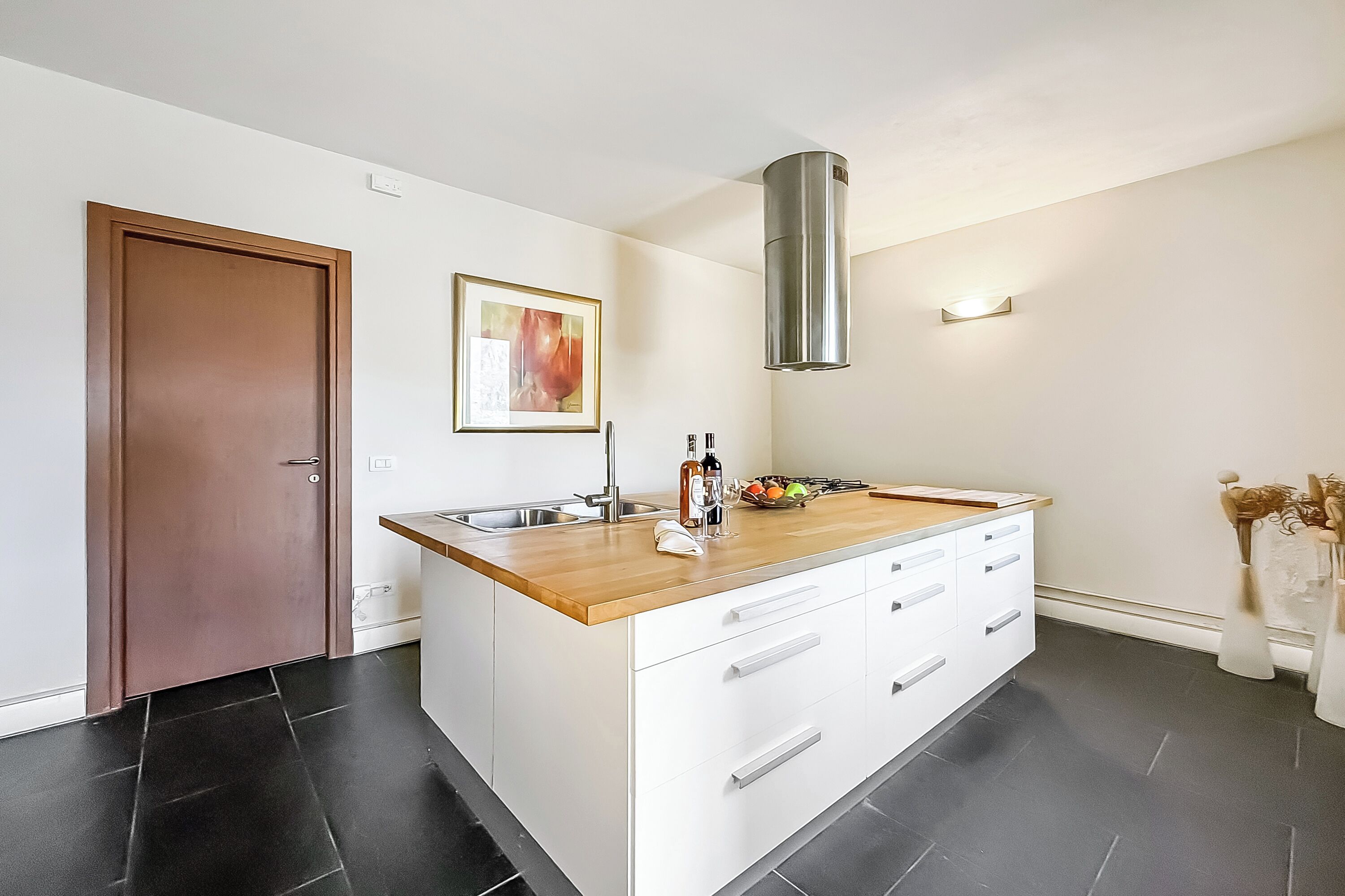 A modern kitchen with a wooden countertop, white cabinets, a stainless steel range hood, and a black tiled floor.