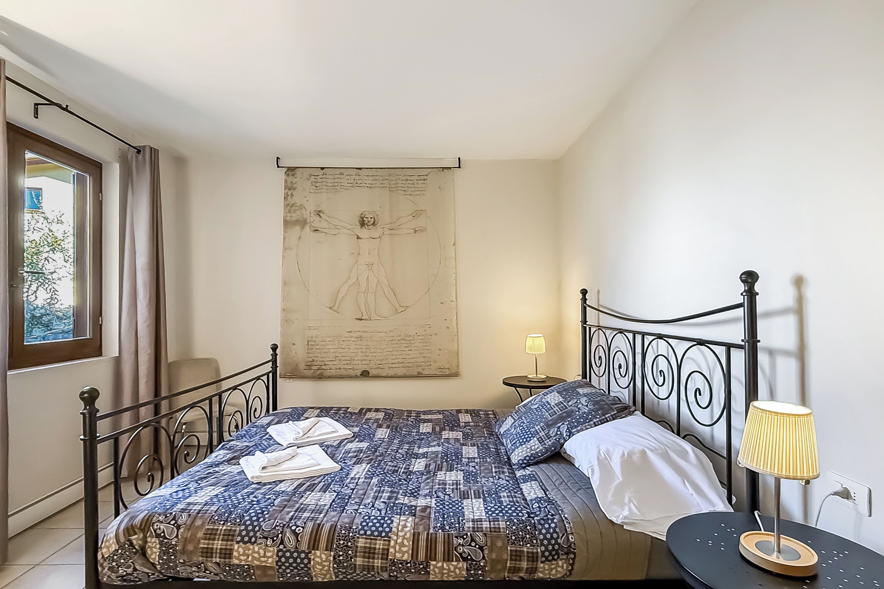 A bedroom with a bed, a bedside table with a lamp, a window with curtains, and a wall with a sketch.