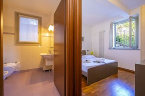 Quarto moderno com cama, janela com vista, banheiro com vaso sanitário e pia, e piso de madeira.