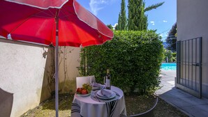 Une table dressée à l’extérieur, avec un parasol rouge, une bouteille de vin et une assiette de raisins.