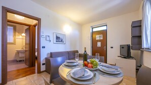 Un coin repas avec une table ronde dressée pour deux personnes, un canapé, un téléviseur et une porte donnant accès à une salle de bain.