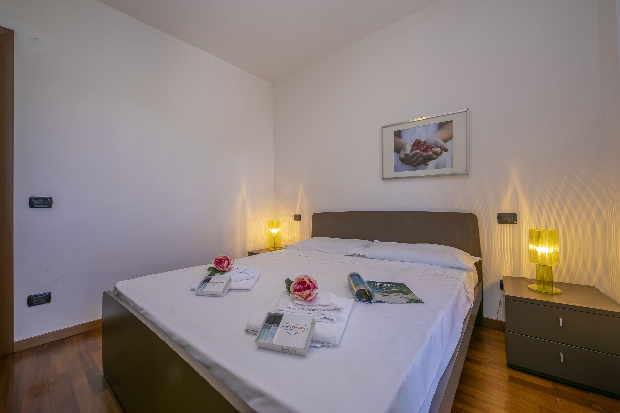 A neatly made bed with a white comforter, two pillows, and a framed picture above it. There are two bedside lamps, a nightstand with a lamp, and a wooden floor.