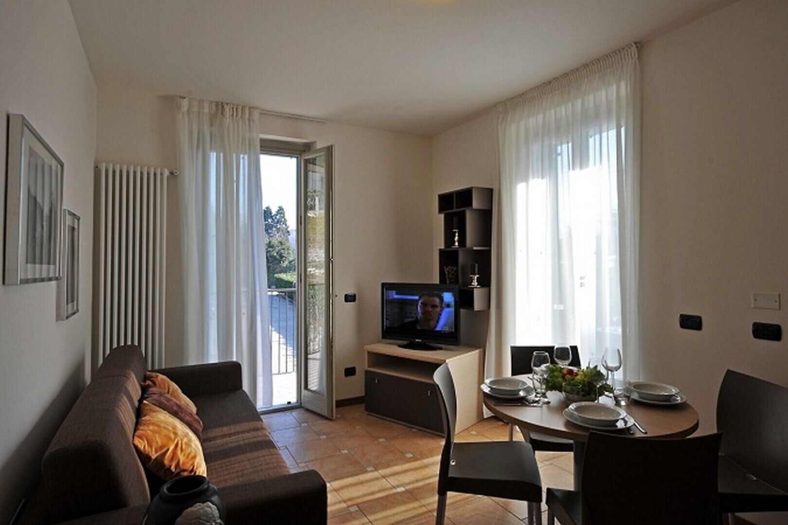 Ett modernt vardagsrum med en soffa, en tv, ett matbord med stolar och en balkong med gardiner.