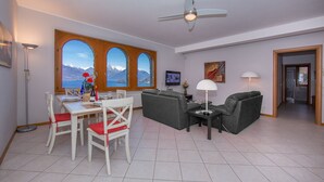 A modern living room with a dining area, large windows with a mountain view, a ceiling fan, and a flat-screen TV. - Casa Monaco (Pianello del Lario)