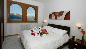 A hotel room with a bed, a window with a view of mountains, a ceiling fan, and a wall with abstract art. - Casa Monaco (Pianello del Lario)