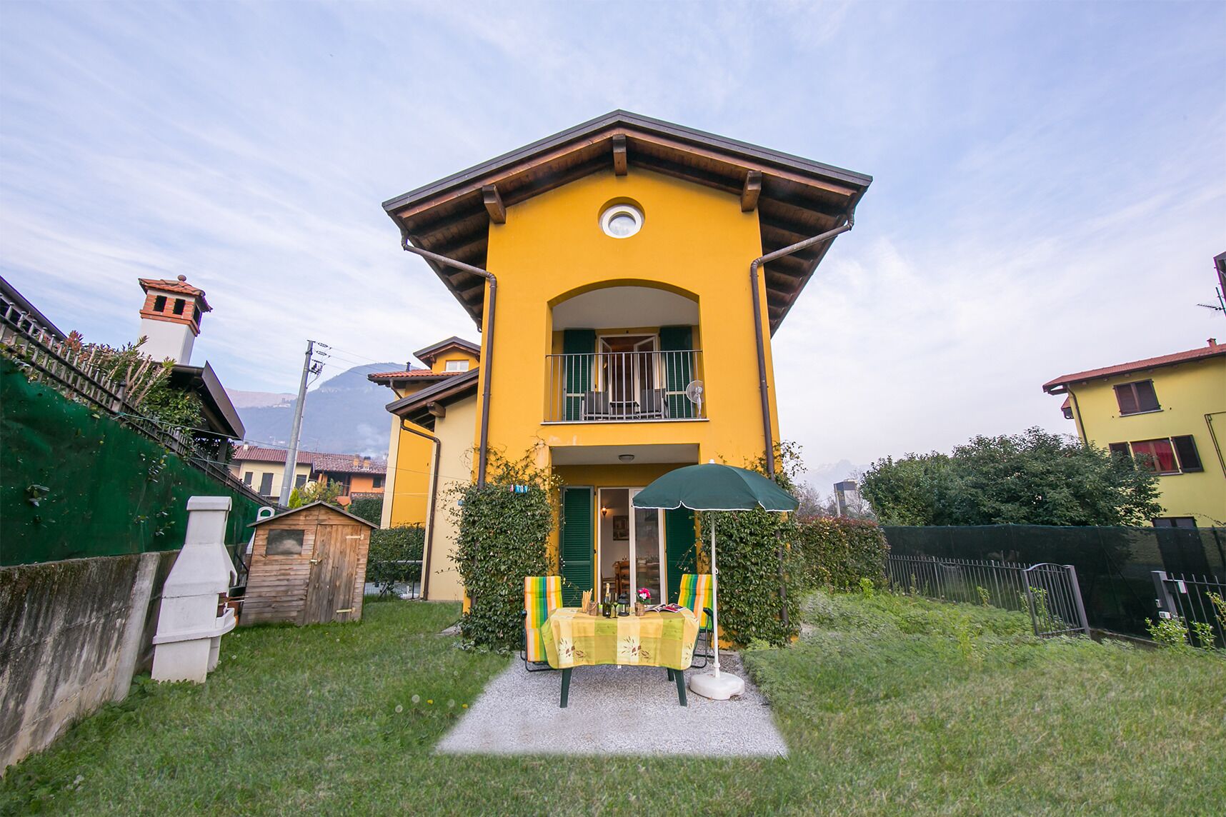 A yellow two-story house with a balcony, a small garden, and a green fence.