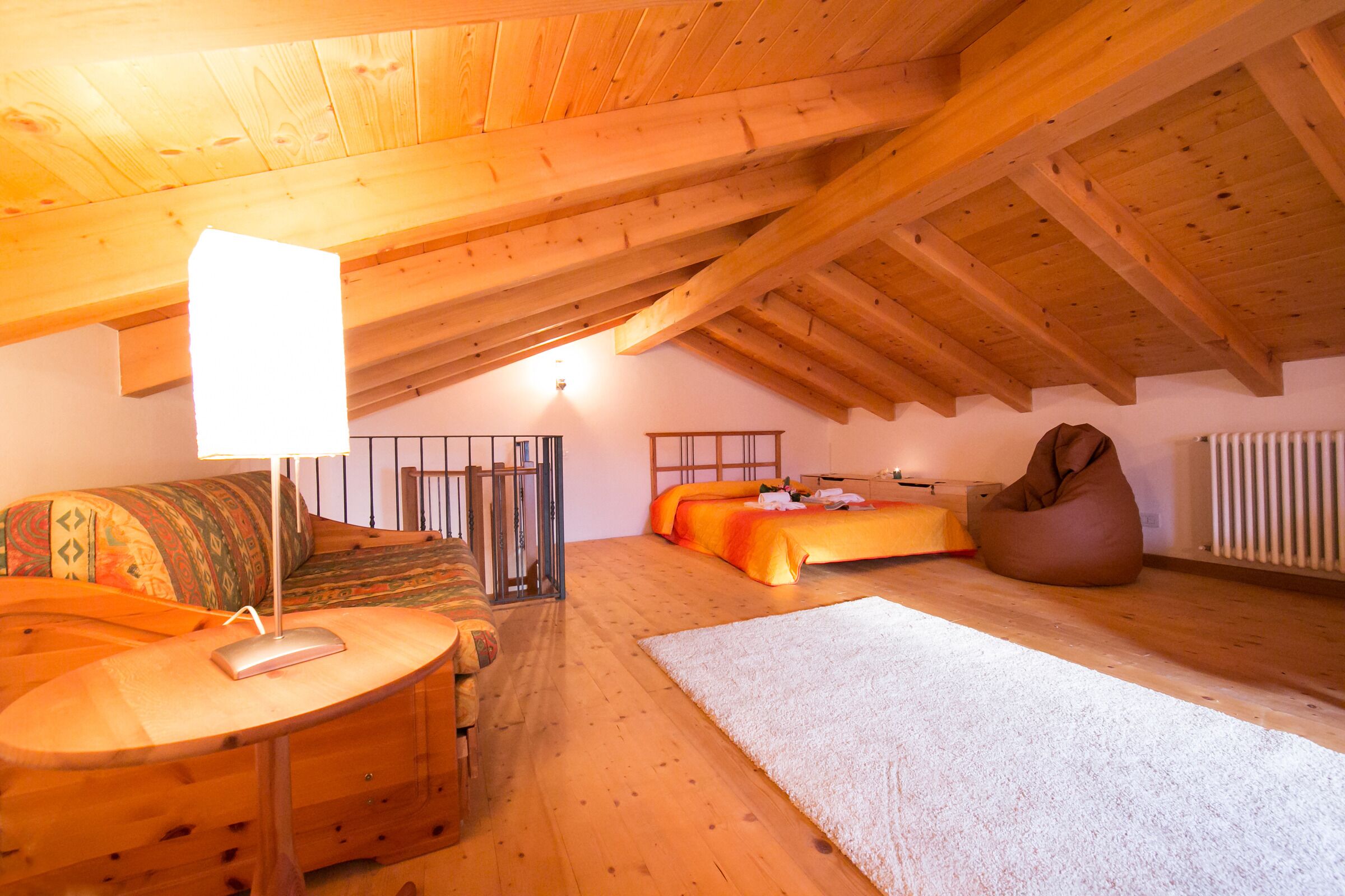 A cozy attic bedroom with a wooden ceiling, a bed, a bean bag, a desk with a lamp, and a radiator.
