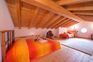 Une chambre avec un plafond en pente, des poutres en bois, un grand lit recouvert d’une couverture colorée, un deuxième lit, une chaise et une fenêtre ronde.