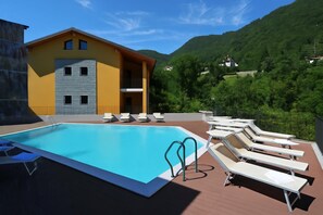 A swimming pool with lounge chairs, a building with a balcony, and a mountainous backdrop. - Residence Eden Magnolia 13 (Argegno)