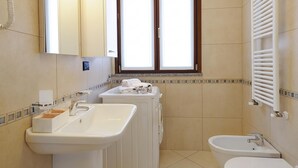 A bathroom with a sink, washing machine, bidet, and a window with blinds. - Residence Eden Magnolia 13 (Argegno)