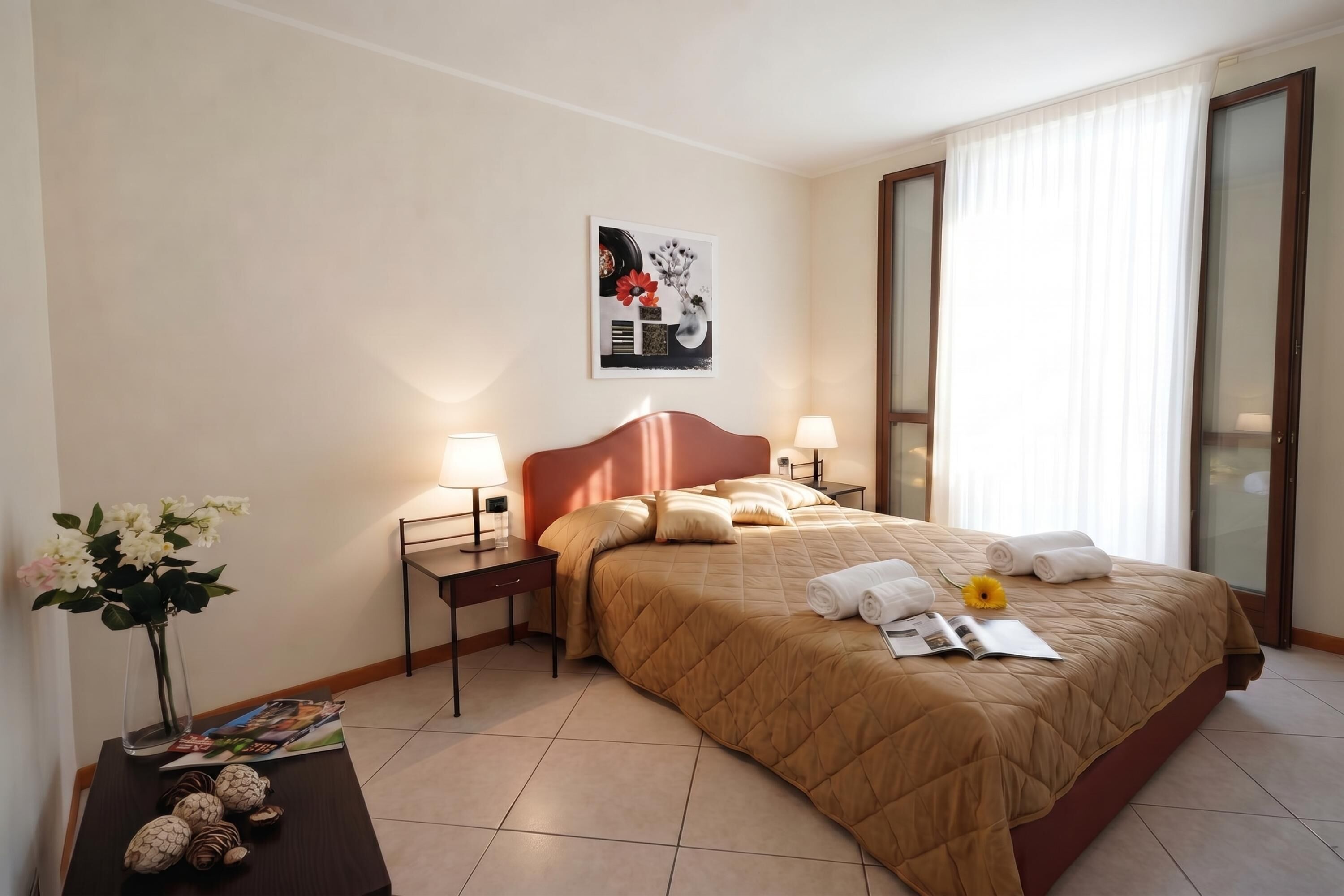 Habitación de hotel con cama, dos mesitas de noche, una lámpara de noche, un florero con flores, una revista y un cuadro en la pared.