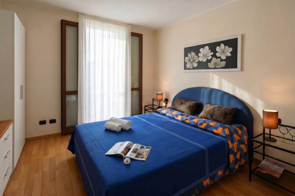 A bedroom with a bed, a nightstand, a lamp, a window with curtains, a door, and a wall with a framed picture.