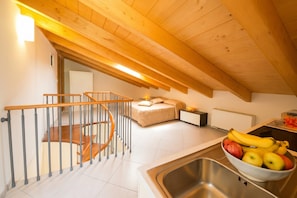 Une cuisine avec un plafond en bois, un escalier doté d’une rampe en métal, un lit et un panier de fruits posé sur le comptoir.