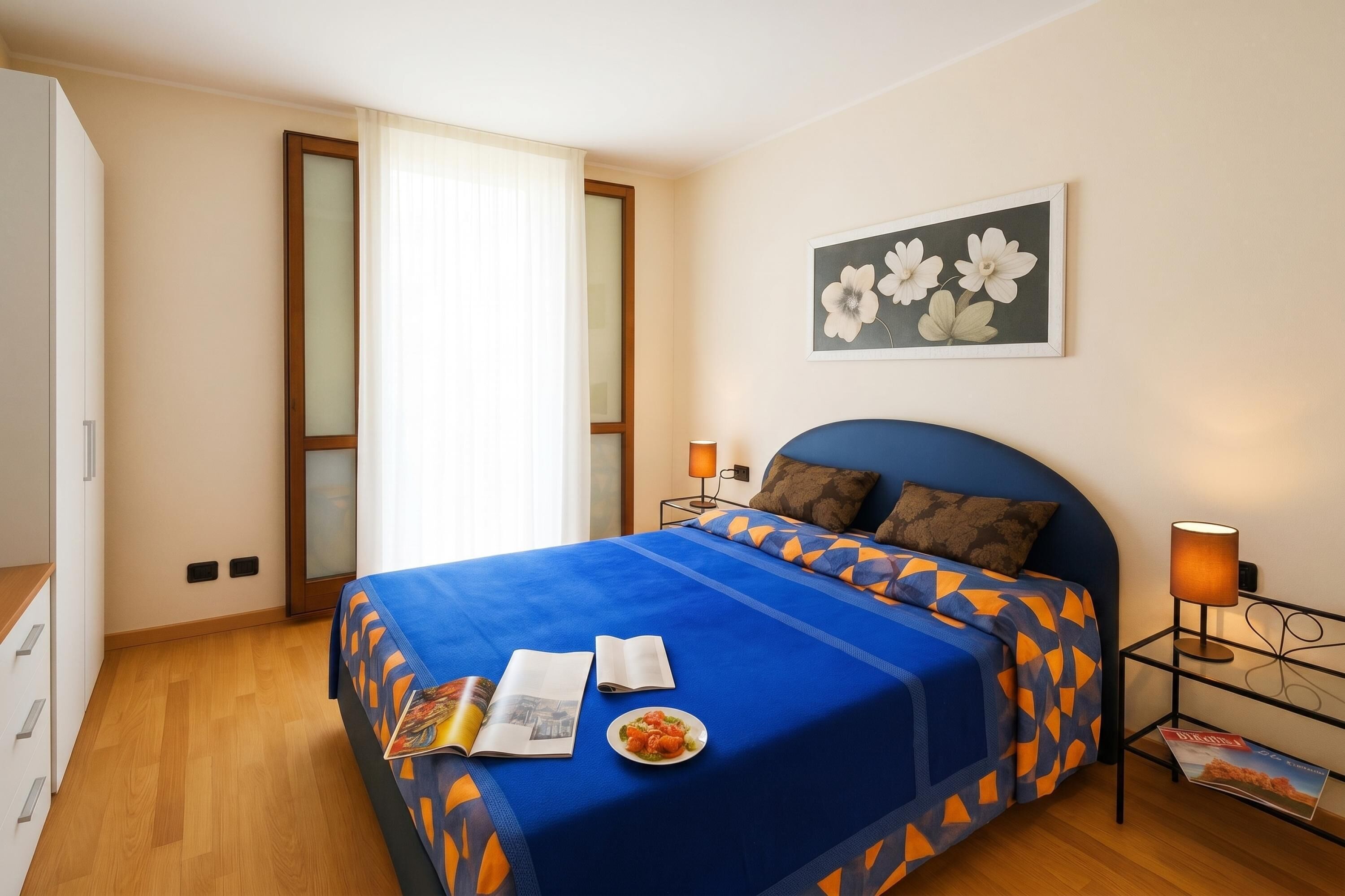 A bedroom with a bed, a nightstand, a lamp, a framed picture, a window with curtains, and a wooden floor.