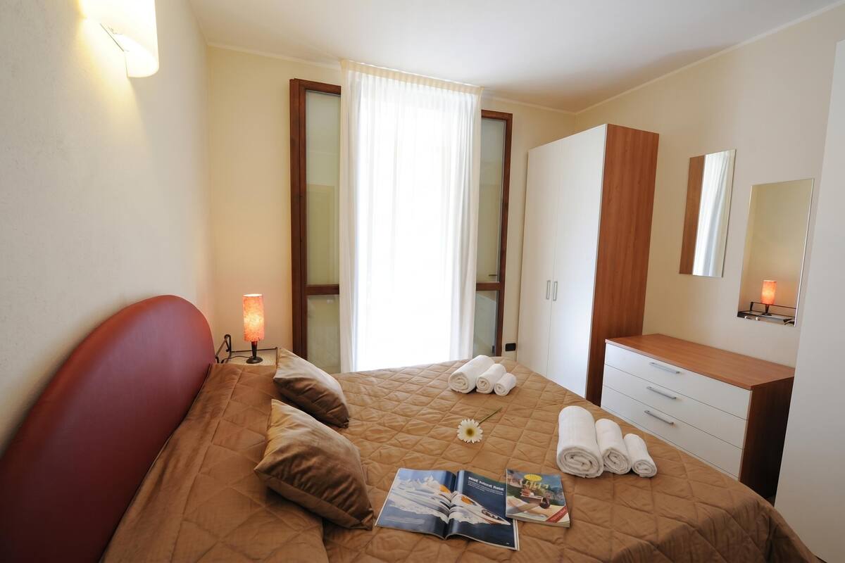 A hotel room with a bed, a nightstand, a lamp, a mirror, a dresser, and a window with curtains.