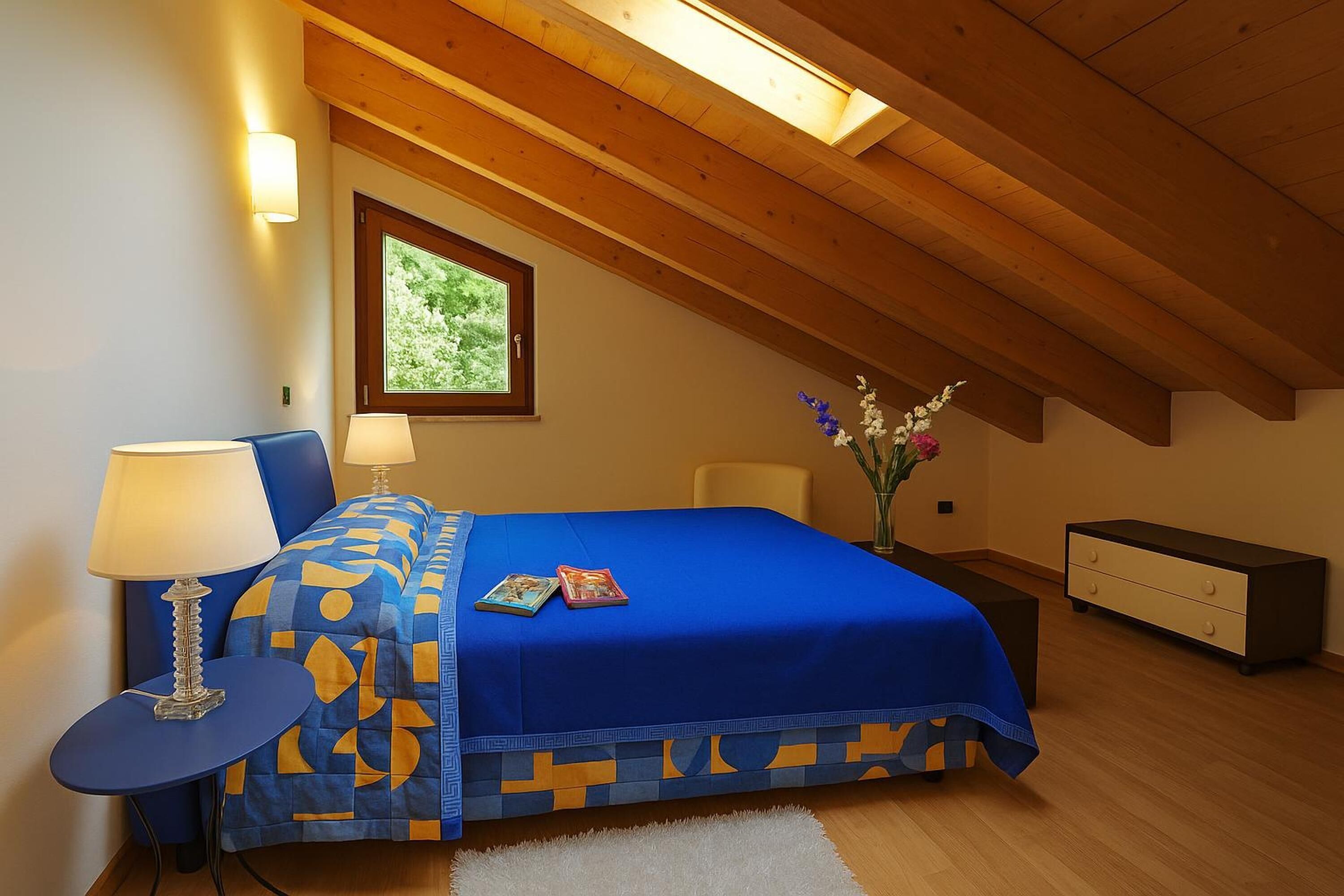 Un dormitorio con techo inclinado, una cama con colcha de diseño azul y amarillo, una mesita de noche con lámpara, un aparador y una ventana con vistas a la vegetación.