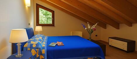 Un dormitorio con techo inclinado, una cama con colcha de diseño azul y amarillo, una mesita de noche con lámpara, un aparador y una ventana con vistas a la vegetación.
