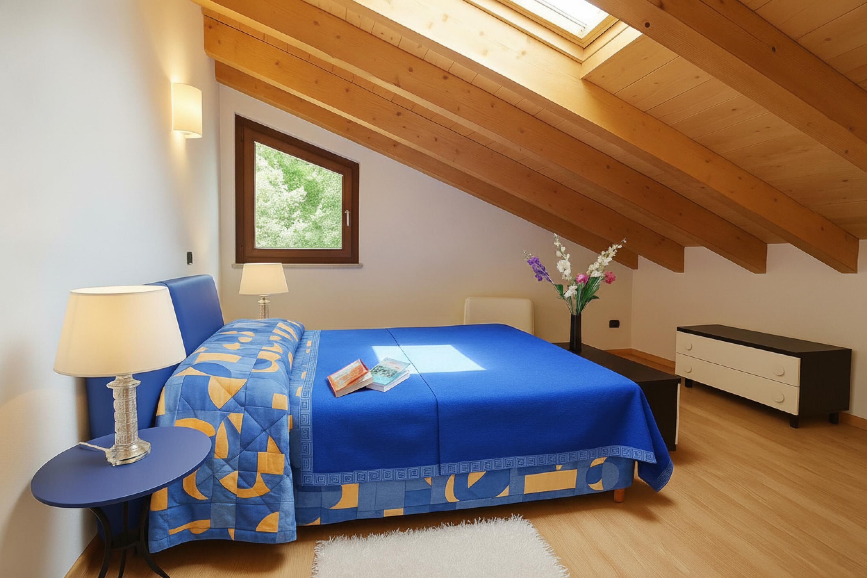 A bedroom with a sloped ceiling, a bed with a blue bedspread, a bedside table with a lamp, a nightstand, and a dresser.